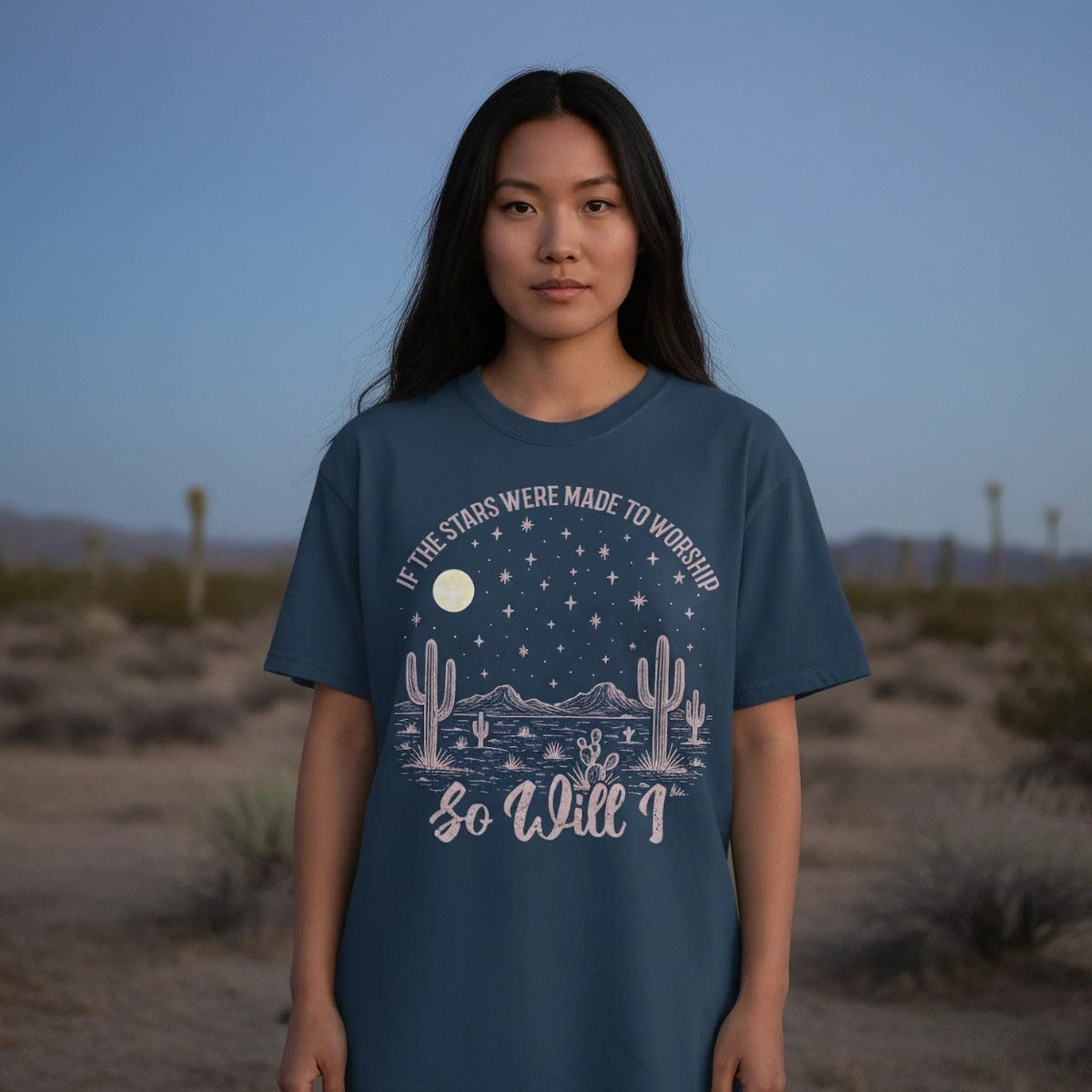 Young woman standing in the desert at dusk wearing a Christian women’s t-shirt that says 'If the stars were made to worship so will I,' featuring moon, stars, mountains, and cactus artwork