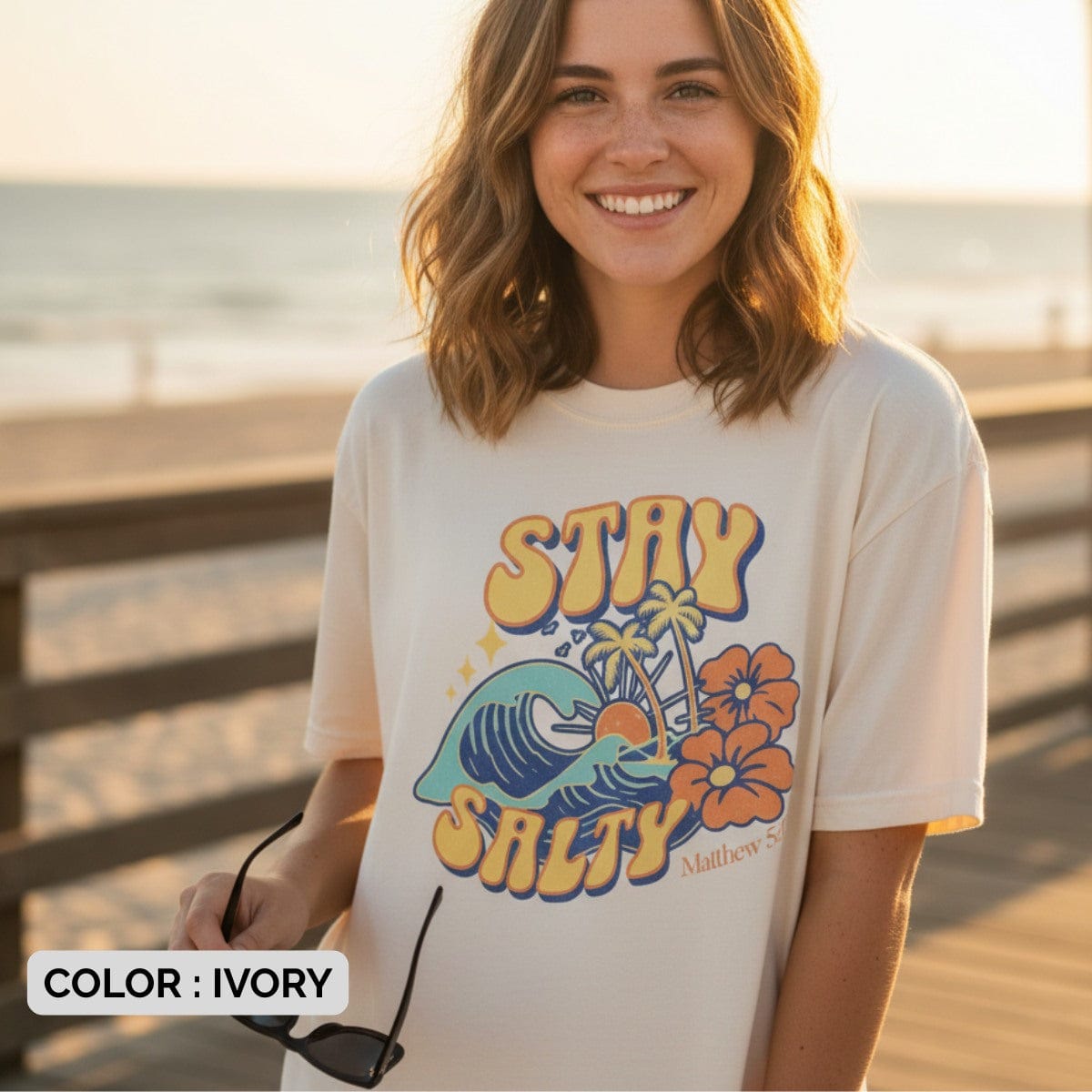 Woman standing on a boardwalk at sunset wearing an Ivory Comfort Colors 1717 tshirt with a retro surf-style design. A summer Christian women tee featuring "Stay Salty" text, waves, palm trees, and Matthew 5:13 scripture, perfect for beachy US faith wear.