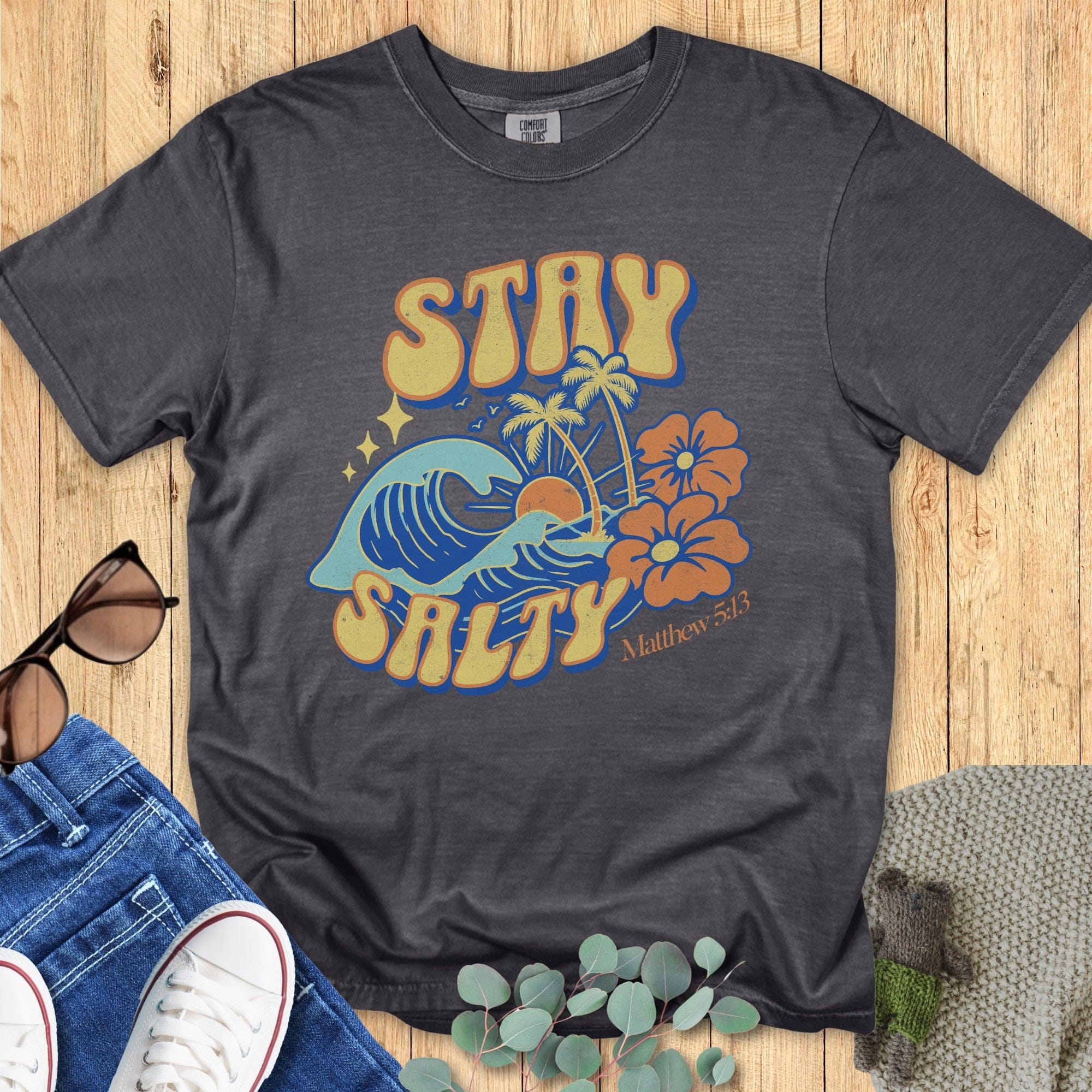 Flatlay of a Comfort Colors 1717 pepper “Stay Salty” t-shirt featuring retro beach graphics with waves, palms, and hibiscus flowers inspired by Matthew 5:13. A fun women’s scripture tee - faith-based apparel for women and modern Christian shirt, great Christian mom gift idea for summer.
