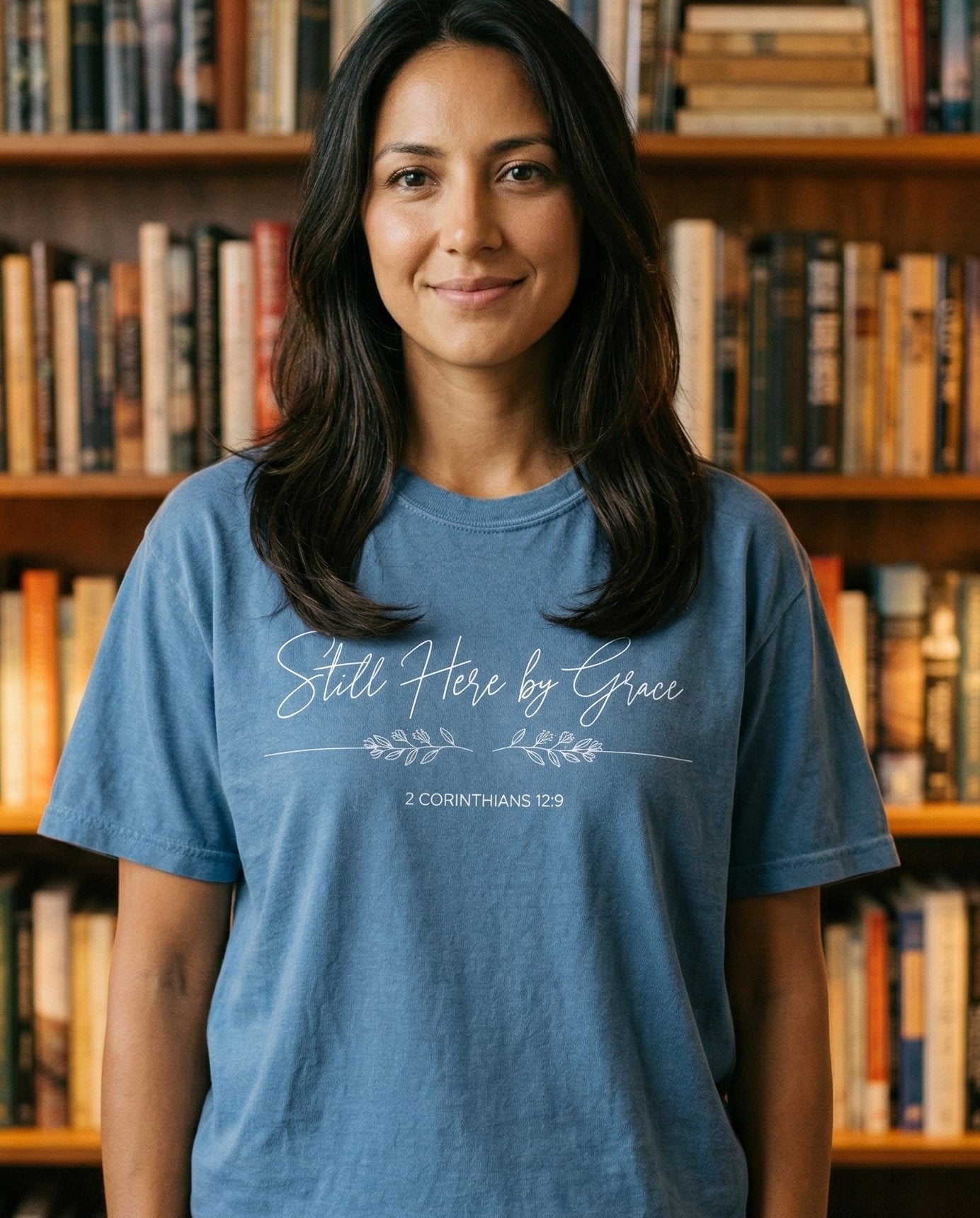 Still Here By Grace – Blue Jean women's Christian tee, lifestyle photo in cozy reading nook – Cursive script reading 'Still Here by Grace' with a delicate leaf bran – 2 Corinthians 12:9 – She Wears Faith