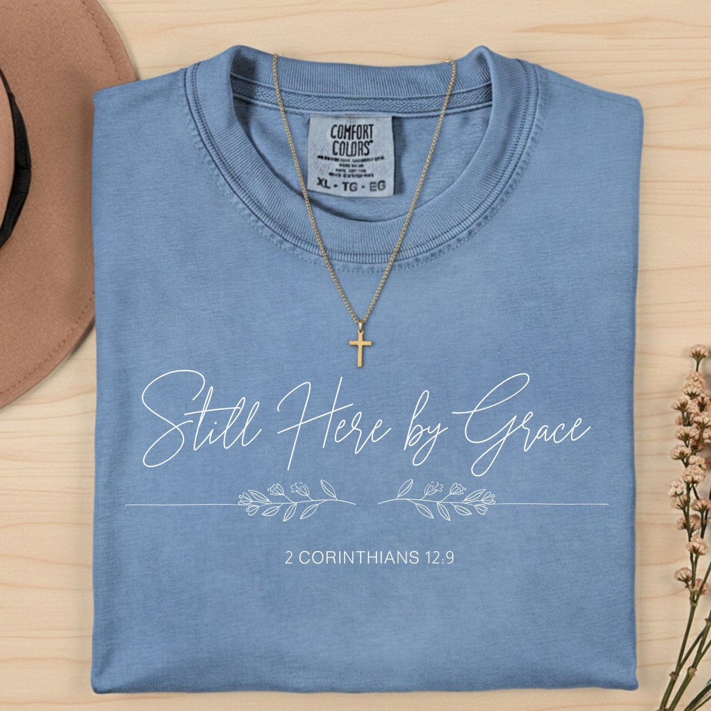 Flatlay of a Comfort Colors 1717 blue jean “Still Here by Grace – 2 Corinthians 12:9” t-shirt with hand-lettered script and floral details. A gentle women’s scripture tee — inspiring faith-based apparel for women and modern Christian shirt, ideal Christian mom gift idea.