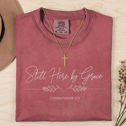 Flatlay of a Comfort Colors 1717 crimson “Still Here by Grace – 2 Corinthians 12:9” t-shirt with minimalist floral line design and delicate lettering. A graceful women’s scripture tee — uplifting faith-based apparel for women and modern Christian shirt, perfect Christian mom gift idea.