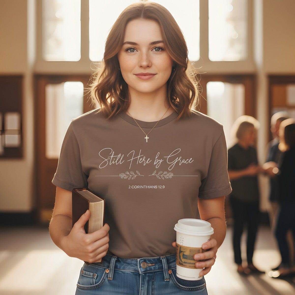 Woman standing in a church lobby holding a Bible and coffee cup, wearing a Comfort Colors 1717 espresso “Still Here by Grace – 2 Corinthians 12:9” t-shirt. A heartfelt women’s scripture tee — faith-based apparel for women and modern Christian shirt, thoughtful Christian mom gift idea.