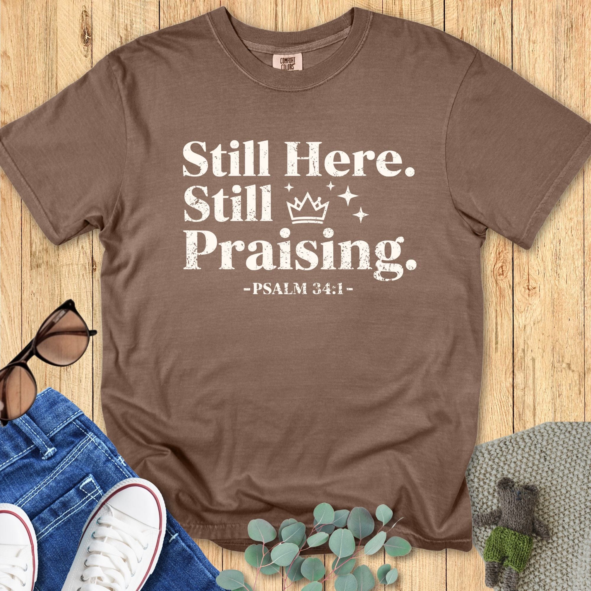 Comfort Colors 1717 Espresso brown Christian T-shirt with the text ‘Still Here. Still Praising. -Psalm 34:1’ in white distressed font, styled with jeans, sunglasses, and sneakers on a wooden background