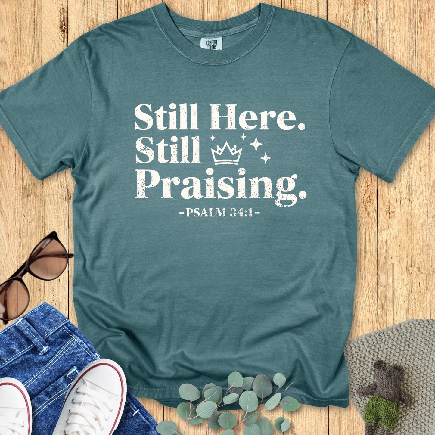 Comfort Colors 1717 Blue Spruce Christian T-shirt featuring the words ‘Still Here. Still Praising. -Psalm 34:1’ in bold white distressed font, styled with jeans, sunglasses, and eucalyptus leaves.