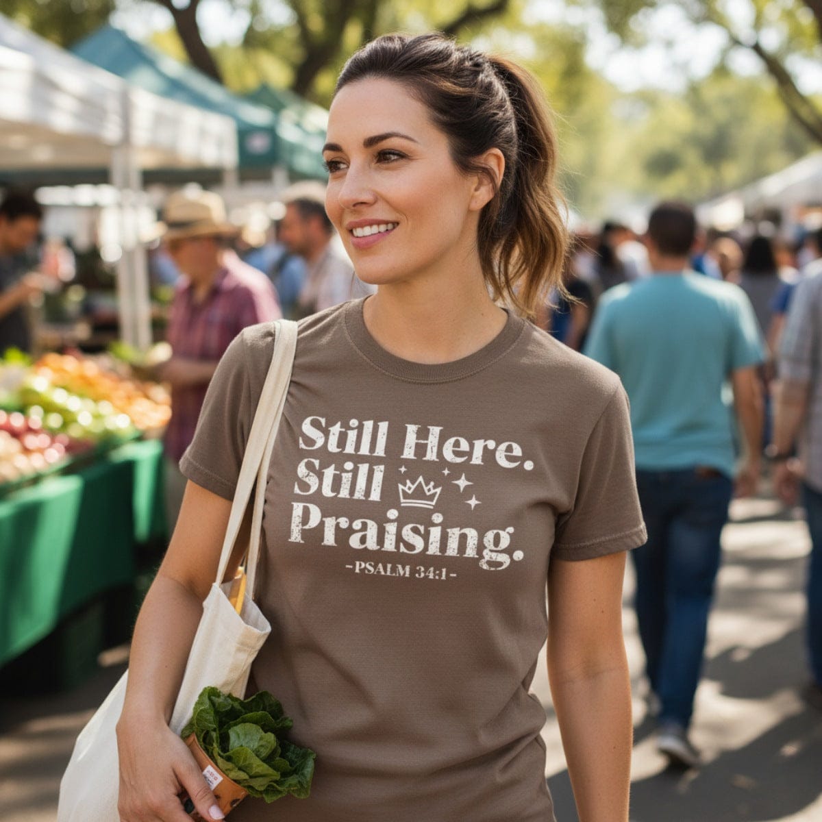 Smiling woman at an outdoor farmer’s market wearing a brown Christian T-shirt with the text ‘Still Here. Still Praising. -Psalm 34:1’ in white distressed font, carrying a tote bag with fresh greens