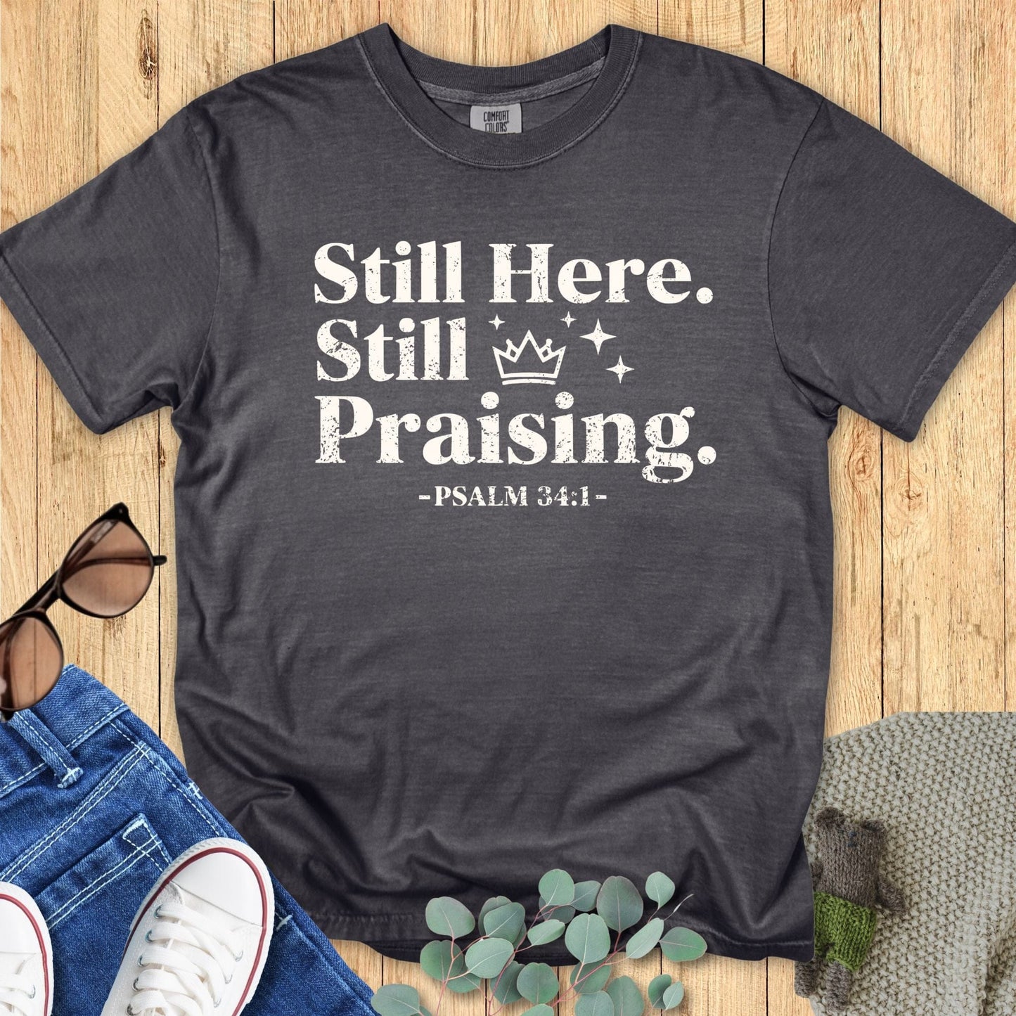 Comfort Colors 1717 Pepper gray Christian T-shirt with the phrase ‘Still Here. Still Praising. -Psalm 34:1’ in white distressed lettering, laid flat with casual accessories.