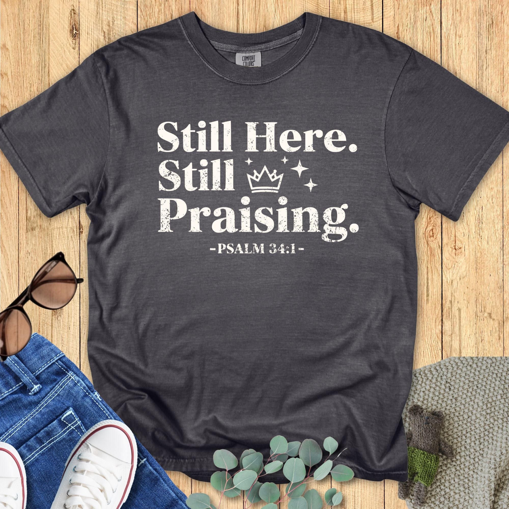 Comfort Colors 1717 Pepper gray Christian T-shirt with the phrase ‘Still Here. Still Praising. -Psalm 34:1’ in white distressed lettering, laid flat with casual accessories.