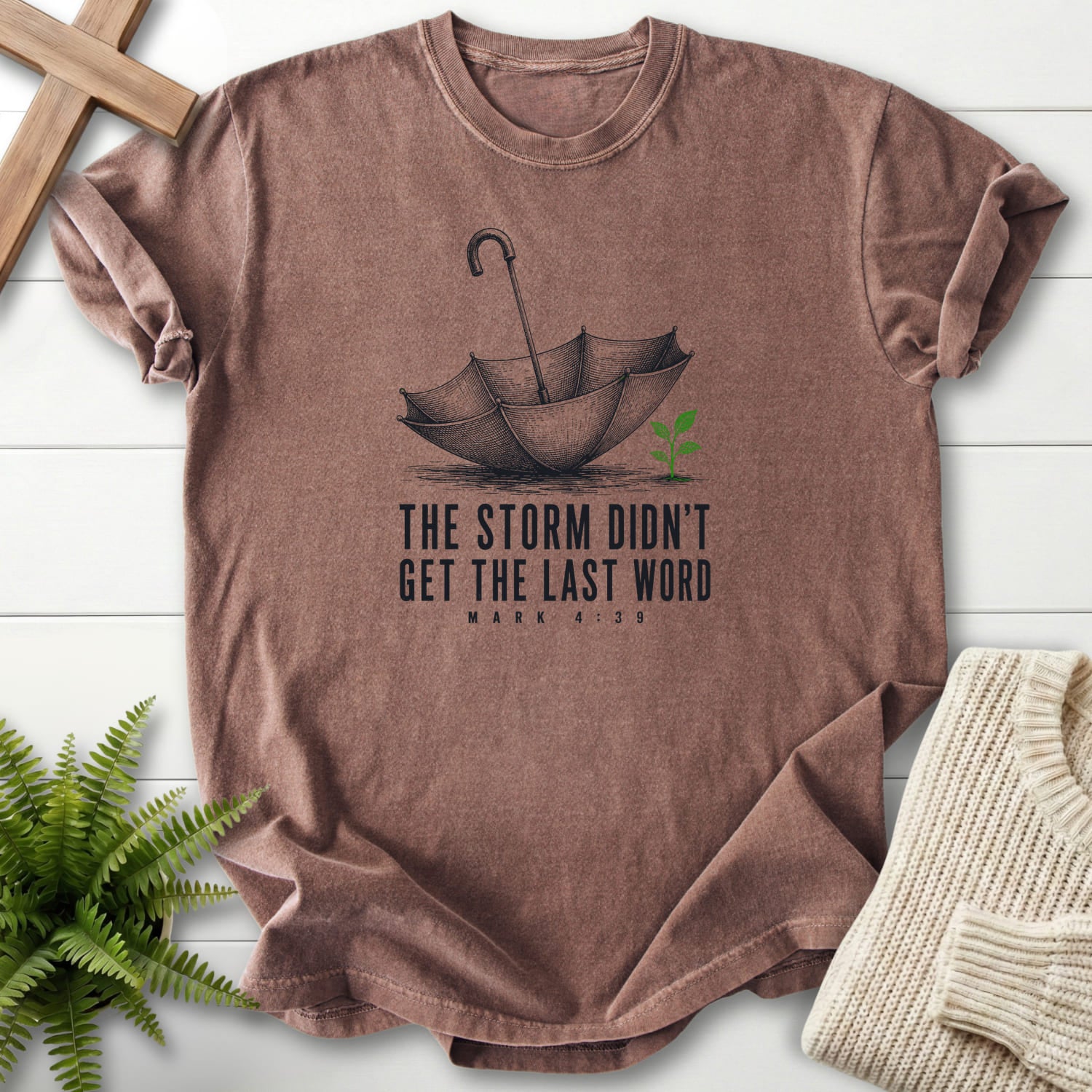 Flat lay of an Espresso women's Christian tee featuring an engraved umbrella and seedling with "Storm Didn't Get the Last Word T-Shirt".