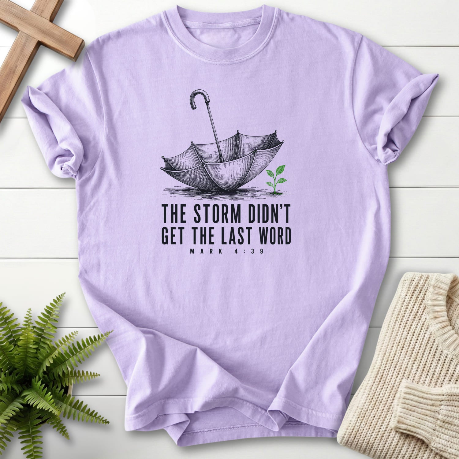 Flat lay of an Orchid Christian t-shirt for women featuring an engraved umbrella and seedling with "Storm Didn't Get the Last Word T-Shirt".