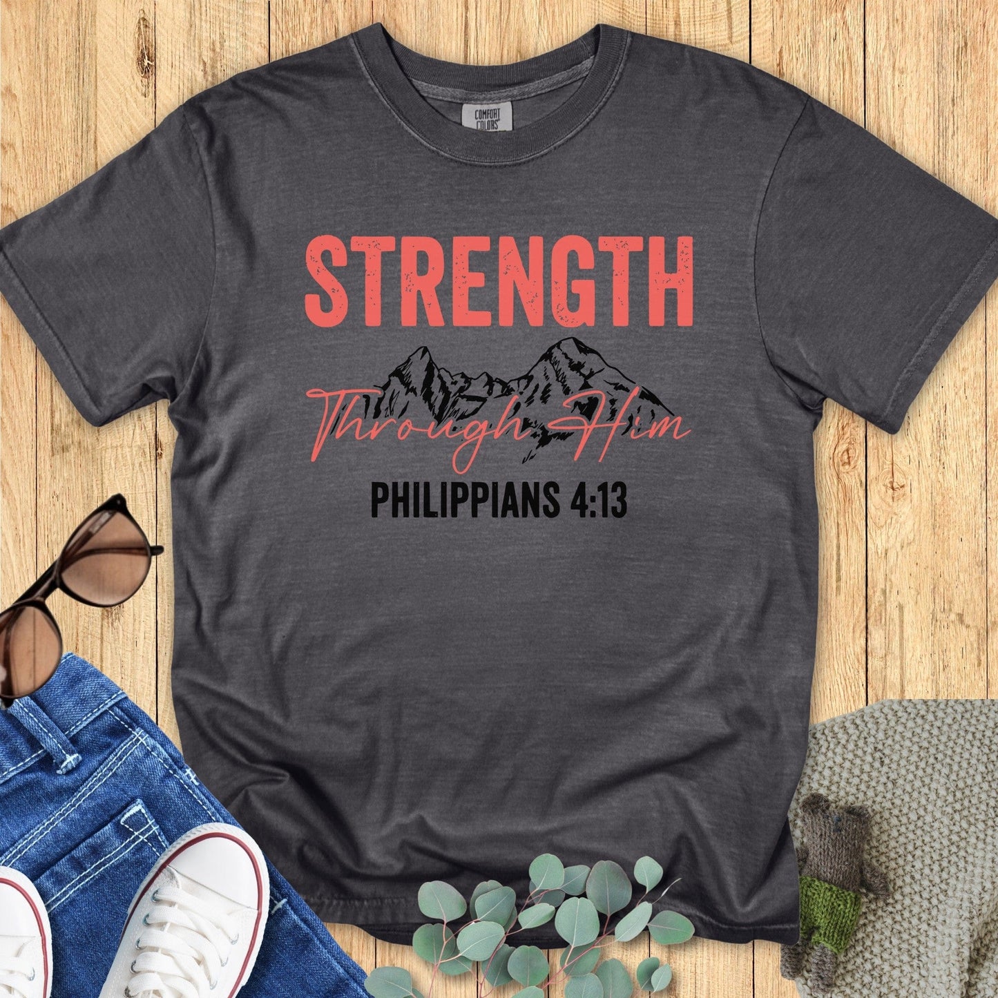Flatlay of a Comfort Colors 1717 pepper “Strength Through Him – Philippians 4:13” t-shirt featuring bold coral typography with mountain artwork. A strong and inspiring women’s scripture tee — modern faith-based apparel for women and meaningful Christian mom gift idea.