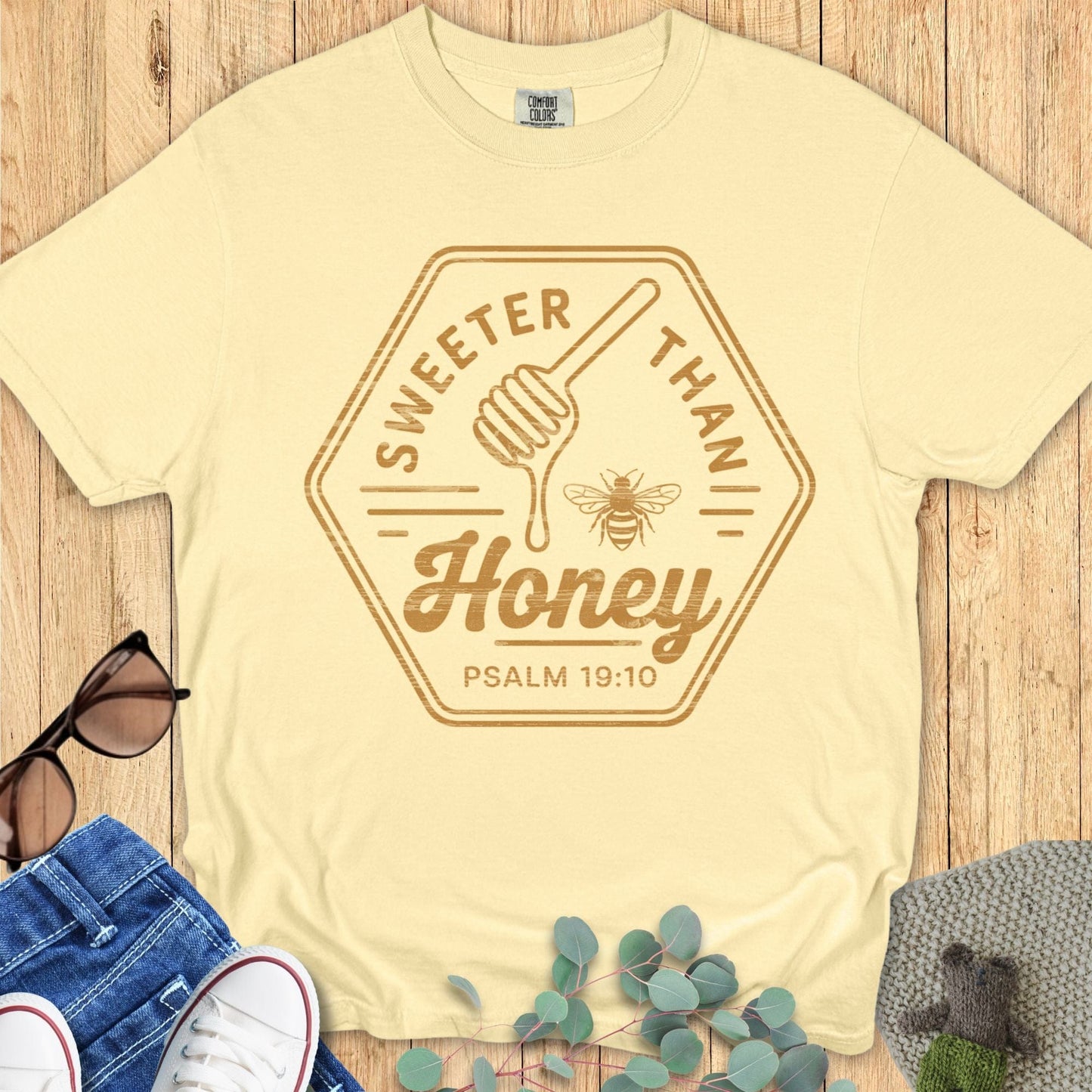 Flat lay of a butter yellow Comfort Colors T-shirt with “Sweeter Than Honey – Psalm 19:10” in golden brown text, honey dipper, and bee graphic. Styled with jeans, sneakers, sunglasses, and greenery on a wood background.