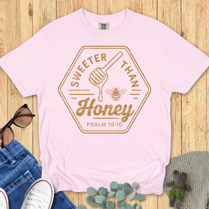 Flat lay of a light pink Comfort Colors T-shirt with “Sweeter Than Honey – Psalm 19:10” in golden brown text, honey dipper, and bee graphic. Styled with jeans, sneakers, sunglasses, and greenery on a wood background