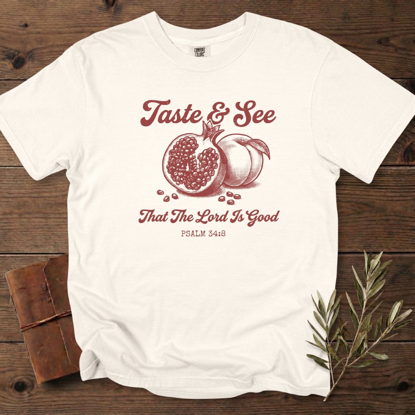 Ivory Christian women tshirt on Comfort Colors 1717 with “Taste & See That The Lord Is Good” Psalm 34:8 design, illustrated with hand-drawn pomegranate and peach; photographed on a warm rustic setup for US faith wear shoppers.