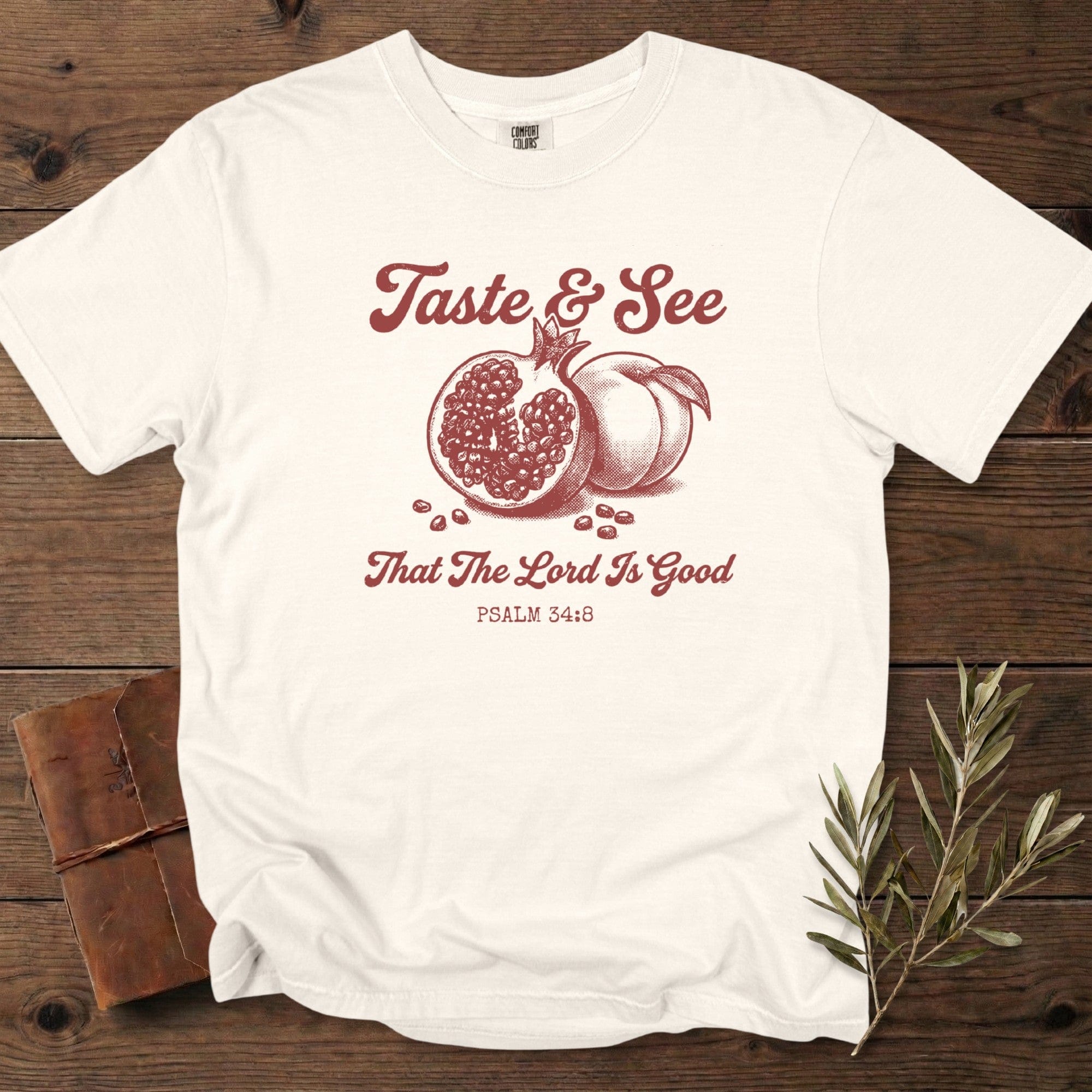 Ivory Christian women tshirt on Comfort Colors 1717 with “Taste & See That The Lord Is Good” Psalm 34:8 design, illustrated with hand-drawn pomegranate and peach; photographed on a warm rustic setup for US faith wear shoppers.