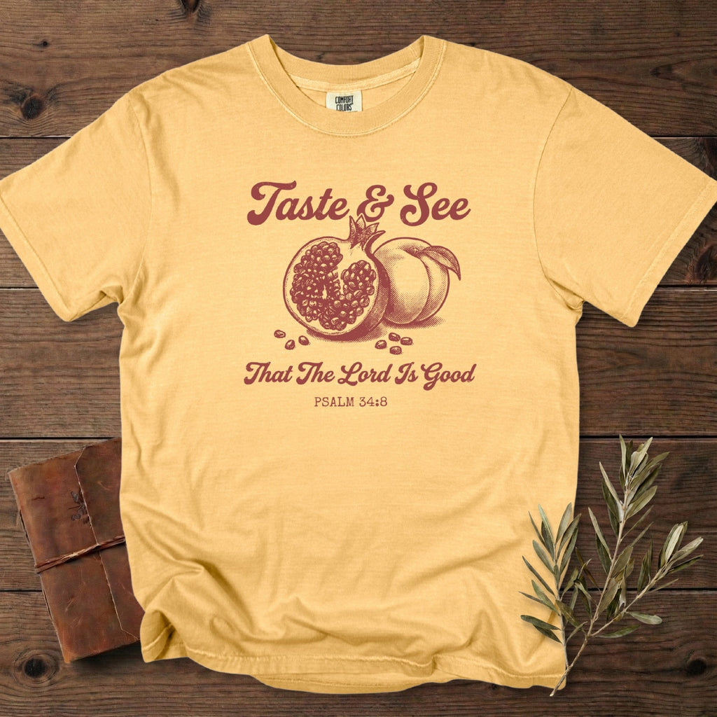 Mustard Comfort Colors 1717 Christian women tee showing a vintage pomegranate and peach graphic with “Taste & See That The Lord Is Good” Psalm 34:8; stylish US faith wear displayed on a cozy wooden background.