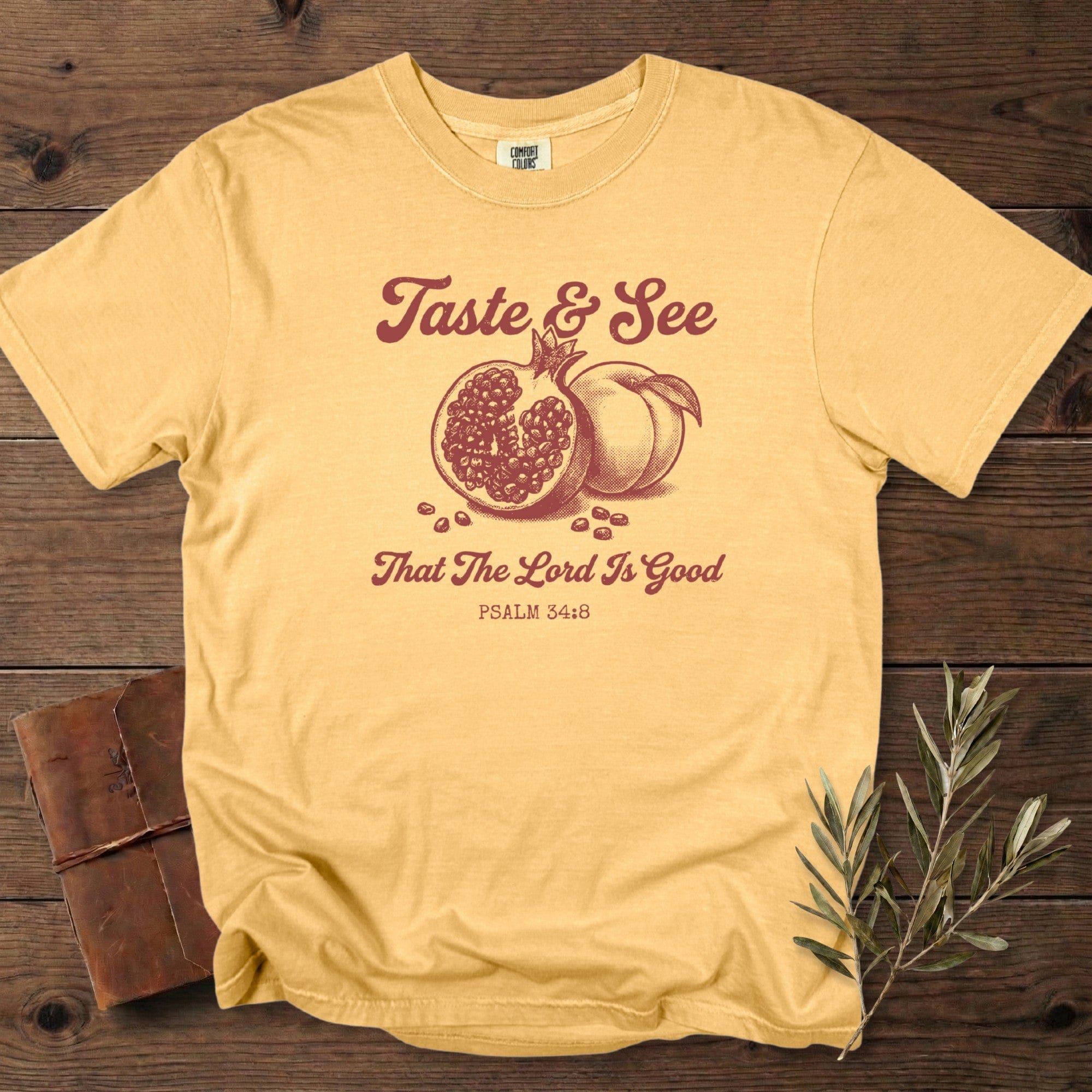 Mustard Comfort Colors 1717 Christian women tee showing a vintage pomegranate and peach graphic with “Taste & See That The Lord Is Good” Psalm 34:8; stylish US faith wear displayed on a cozy wooden background.