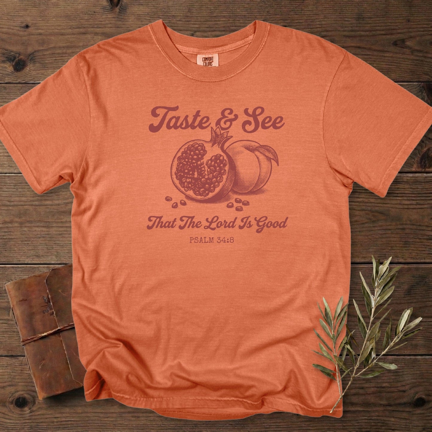 Christian women tee in terracotta Comfort Colors 1717 featuring “Taste & See That The Lord Is Good” from Psalm 34:8 with vintage pomegranate and peach illustration; inspirational US faith wear displayed on a rustic wood background.