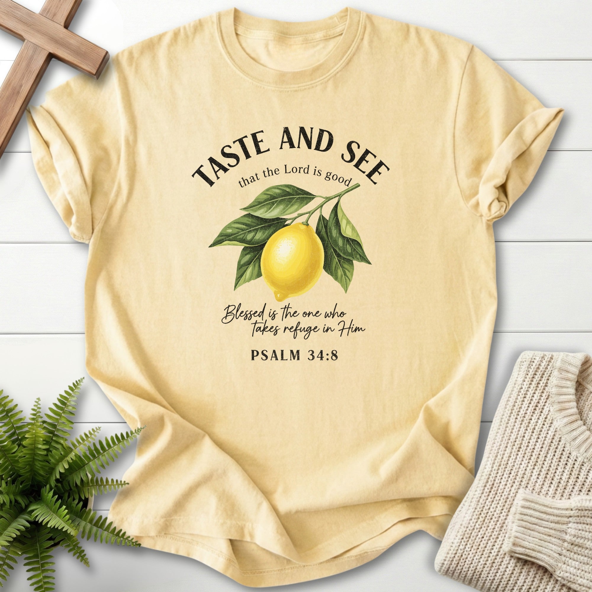 Beige t-shirt with lemon graphic and text on a wooden surface with a plant and sweater.