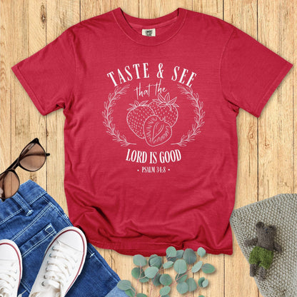 Chili red Comfort Colors Christian T-shirt featuring hand-drawn strawberry and wreath artwork with verse "Taste & See That the Lord Is Good, Psalm 34:8." Faith-based inspirational tee flat lay with jeans, sneakers, and sunglasses on wood background.