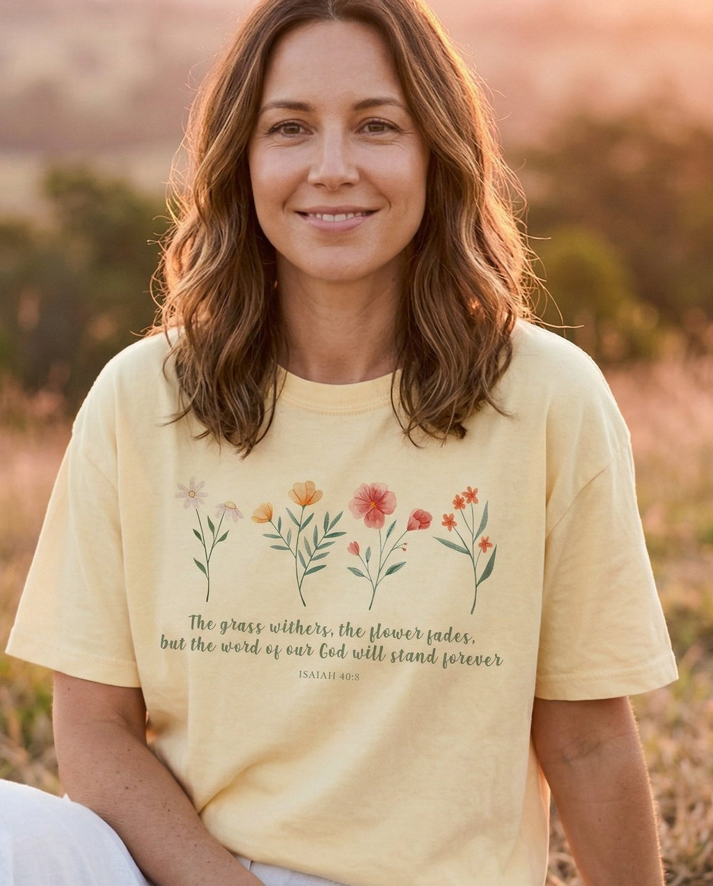 The Grass Withers, The Flower Fades – Banana women's Christian tee, lifestyle photo in wildflower meadow at sunrise – Watercolor wildflower row in soft pink – Isaiah 40:8 – She Wears Faith