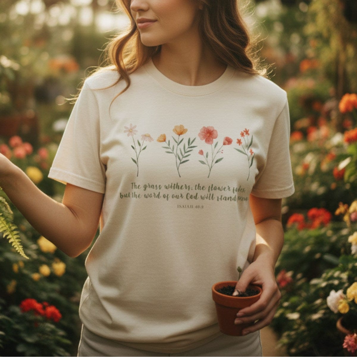 Woman tending plants in a greenhouse wearing a Comfort Colors 1717 ivory “The Grass Withers, The Flower Fades – Isaiah 40:8” t-shirt. A peaceful and elegant women’s scripture tee, making a thoughtful Christian mom gift idea.