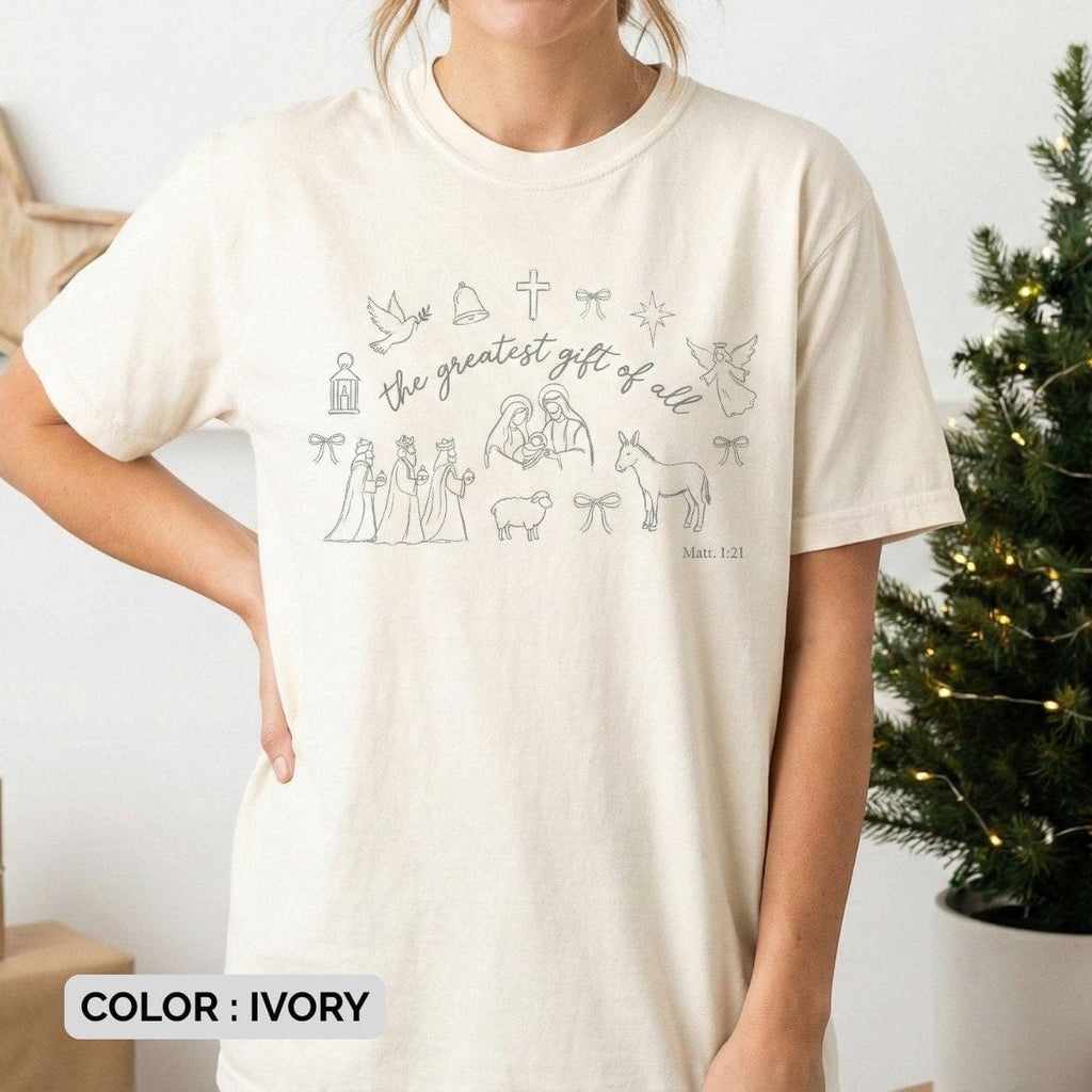 Model wearing an ivory Comfort Colors 1717 Christian women tee featuring minimalist Nativity line art and “the greatest gift of all” Matthew 1:21 design, soft neutral US faith wear styled in a cozy holiday setting.