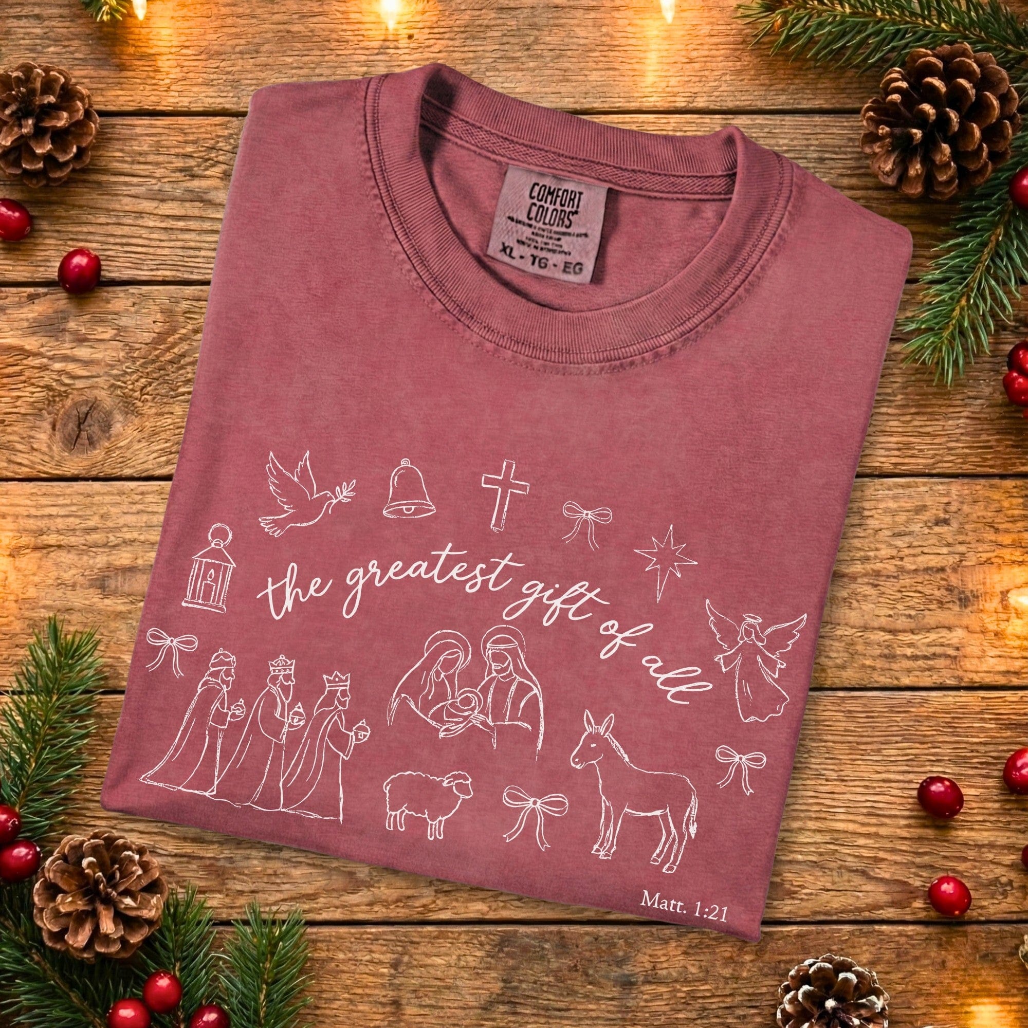 Folded crimson Comfort Colors 1717 Christian women tshirt displaying Nativity scene line art and “the greatest gift of all” scripture design, festive yet minimalist US faith wear on wood background.