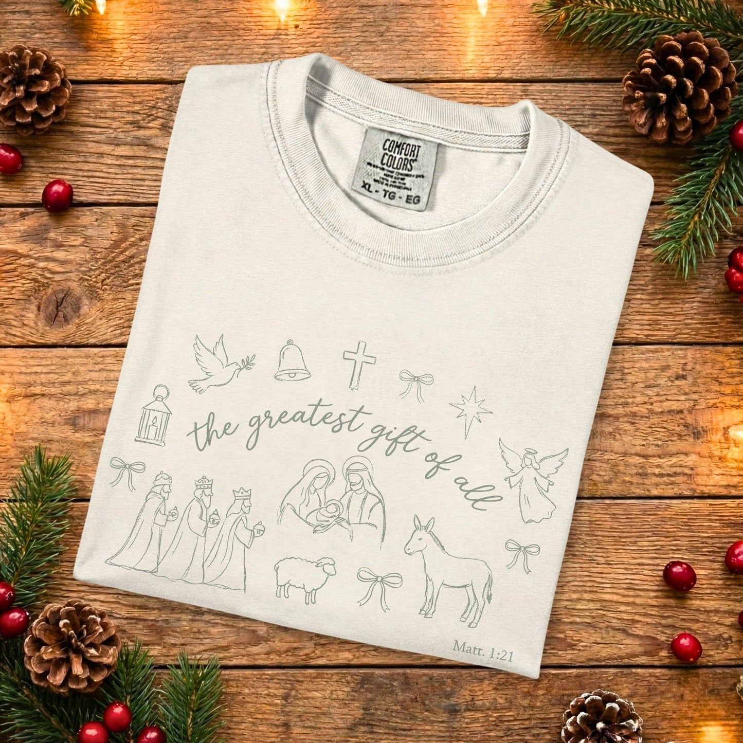 Folded ivory Comfort Colors 1717 Christian women t-shirt featuring minimalist Nativity line art and the phrase “the greatest gift of all” from Matthew 1:21, laid on a rustic wooden background, cozy US faith wear style.
