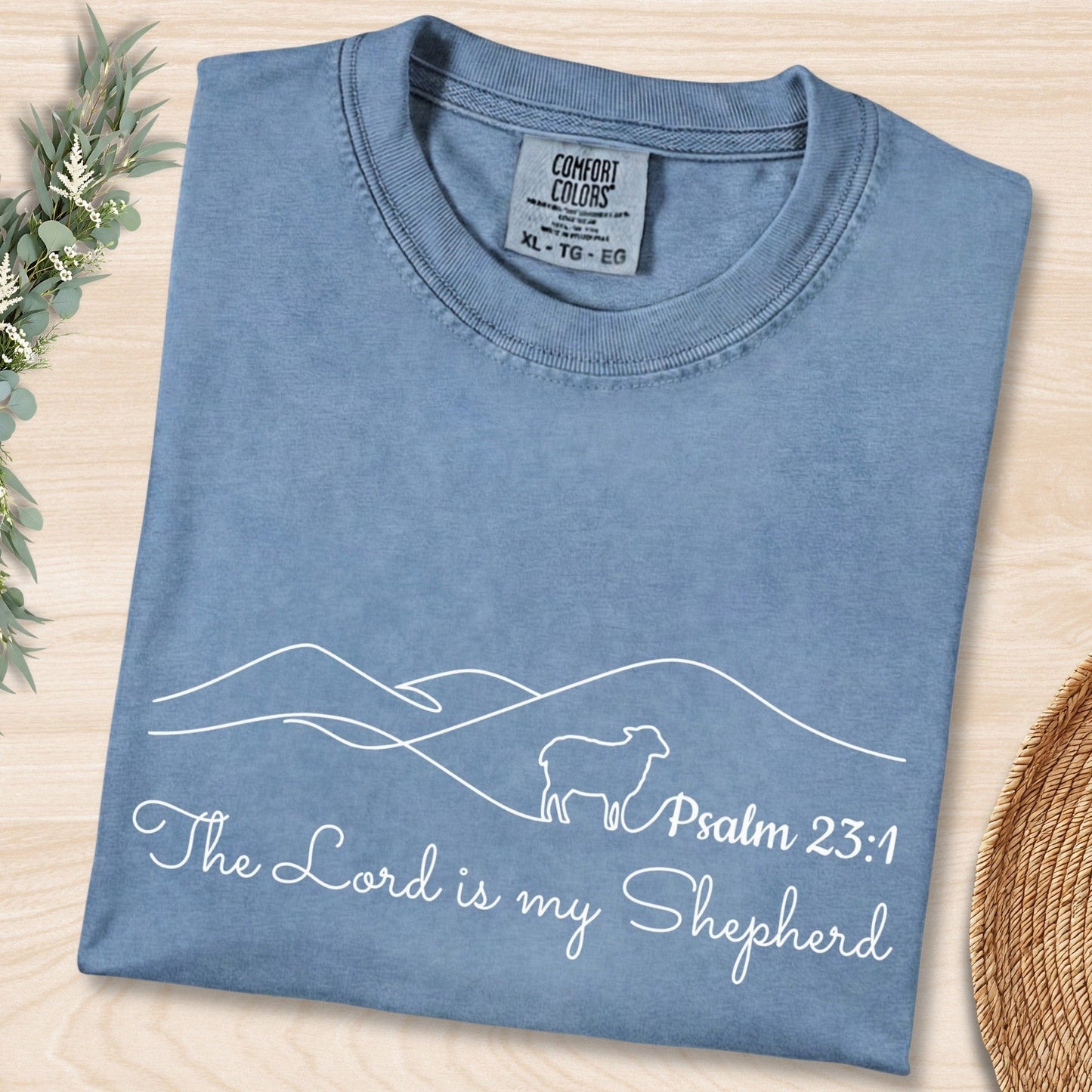 Flat lay of a Blue Jean Comfort Colors 1717 tshirt featuring minimalist white line art of a sheep and mountains. A serene Christian women tee with "The Lord is my Shepherd" Psalm 23:1 scripture, perfect for US faith wear shops.