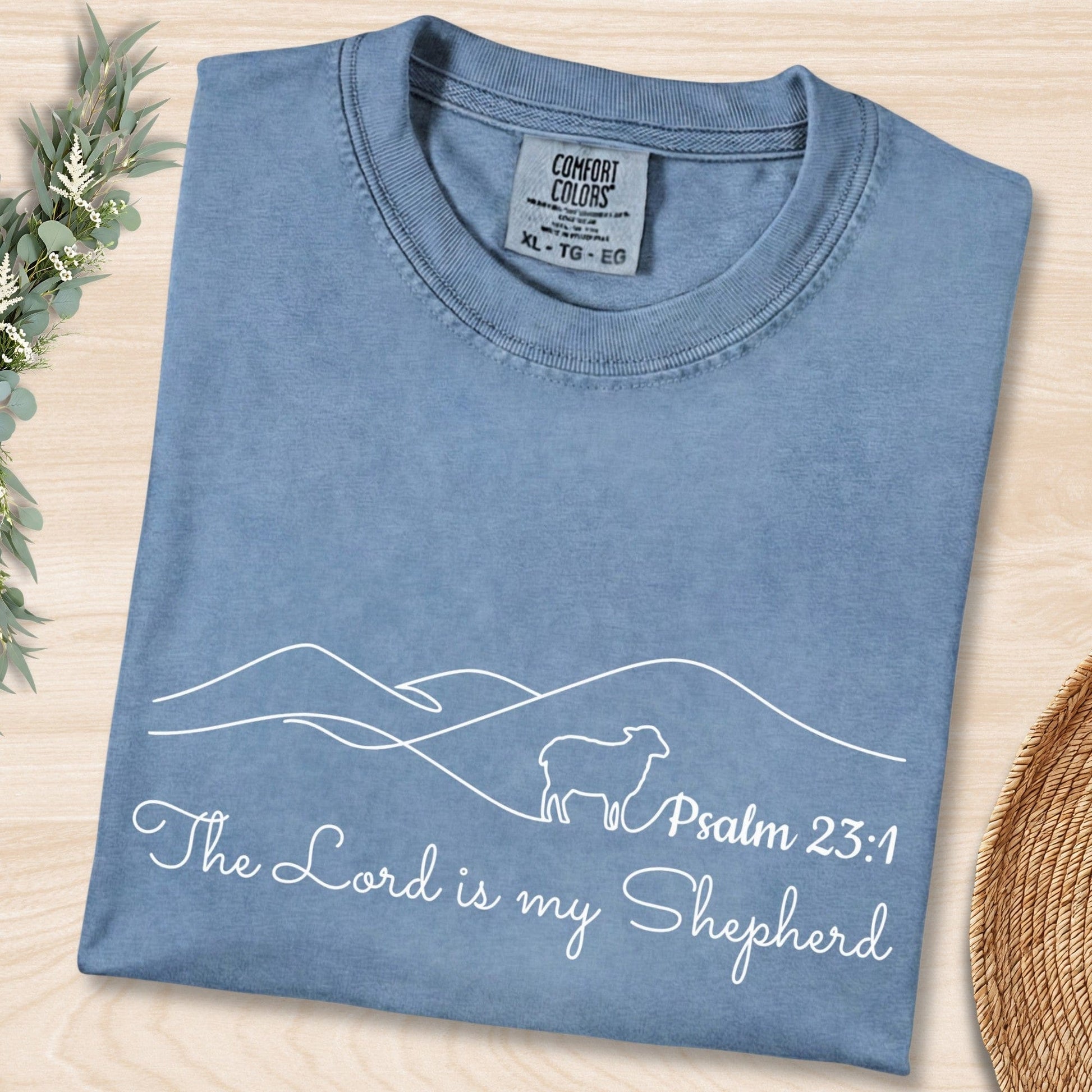 Flat lay of a Blue Jean Comfort Colors 1717 tshirt featuring minimalist white line art of a sheep and mountains. A serene Christian women tee with "The Lord is my Shepherd" Psalm 23:1 scripture, perfect for US faith wear shops.