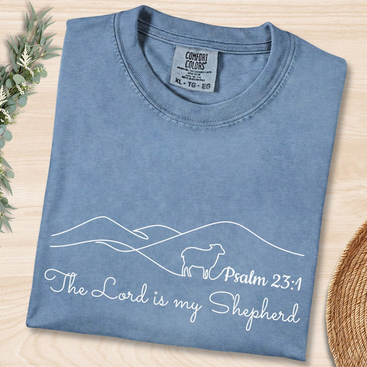 Flat lay of a Blue Jean Comfort Colors 1717 tshirt featuring minimalist white line art of a sheep and mountains. A serene Christian women tee with "The Lord is my Shepherd" Psalm 23:1 scripture, perfect for US faith wear shops.