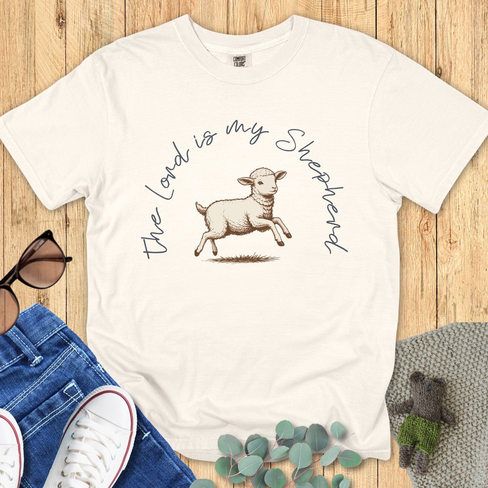 Flatlay of a Comfort Colors 1717 ivory “The Lord Is My Shepherd” t-shirt featuring a hand-drawn lamb illustration. A soft and timeless women’s scripture tee, perfect for Christian mom gift ideas and daily faith wear.