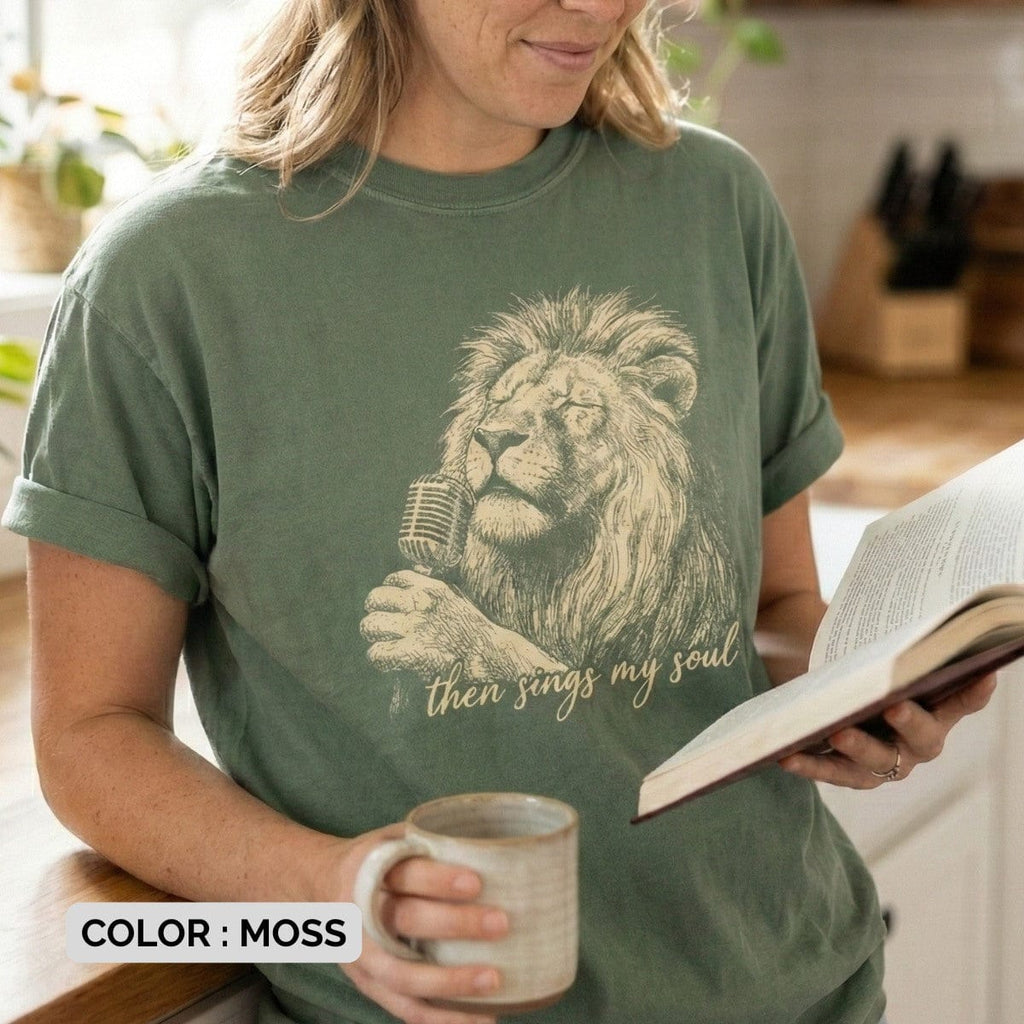 Woman reading a book while wearing a Moss Green Comfort Colors 1717 tshirt with a vintage lion singing graphic. A cozy Christian women tee featuring the lyric "Then Sings My Soul," perfect for US faith wear and worship.