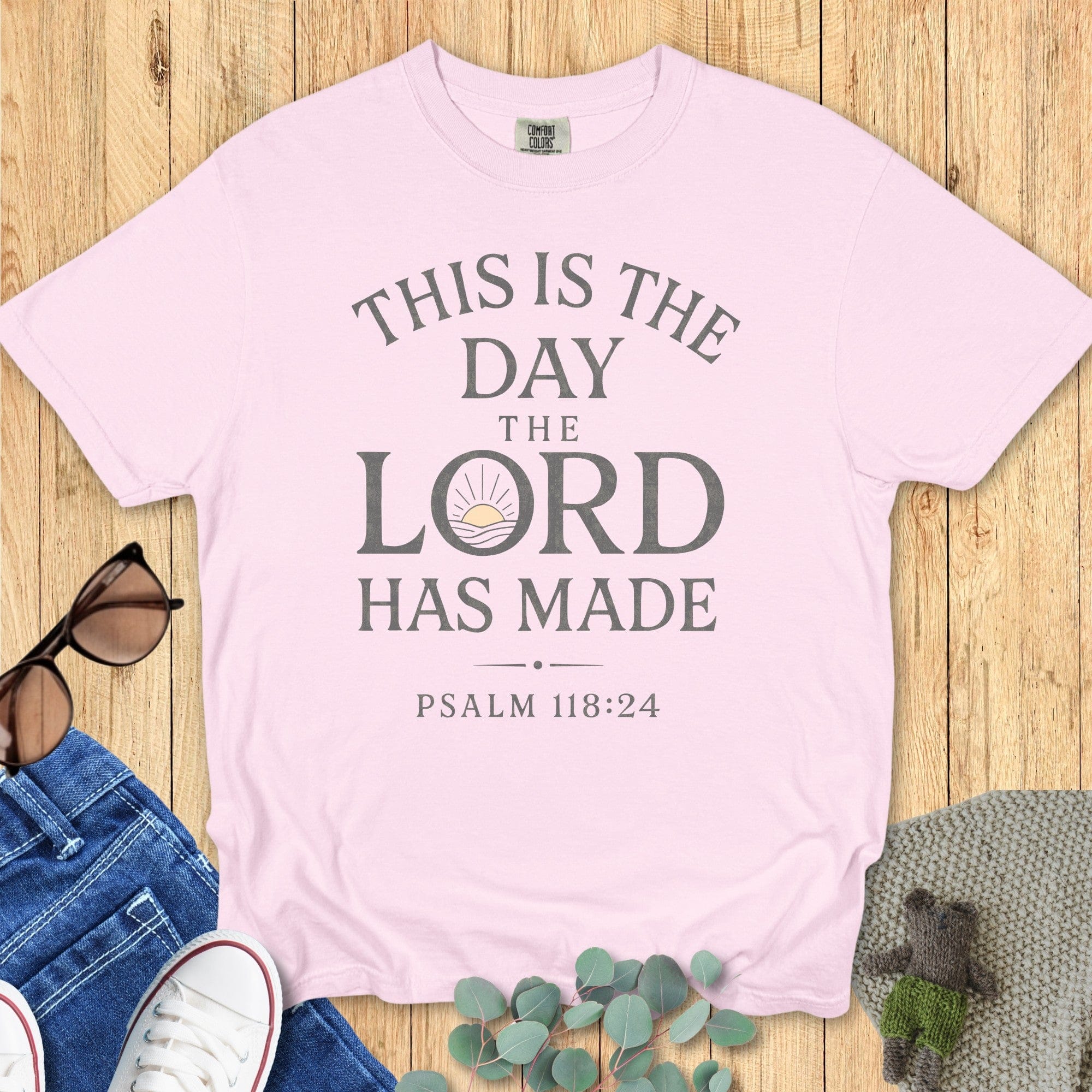Flatlay of a Comfort Colors 1717 blossom pink “This Is The Day The Lord Has Made – Psalm 118:24” t-shirt featuring soft typography and sunrise motif. A cheerful women’s scripture tee and uplifting Christian mom gift idea.