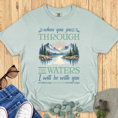 Comfort Colors Christian T-shirt in bay blue with Isaiah 43:2 Bible verse. Design features mountains, forest, and water reflecting the sunrise with text “When you pass through the waters I will be with you.” Flat lay styling includes jeans, white sneakers, sunglasses, and eucalyptus leaves