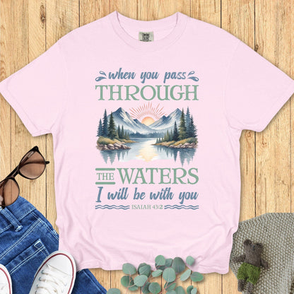 Comfort Colors Christian T-shirt in blossom pink featuring Isaiah 43:2 scripture. Illustrated graphic of mountains and water with sunrise, paired with the words “When you pass through the waters I will be with you.” Displayed flat with casual jeans, shoes, and accessories.