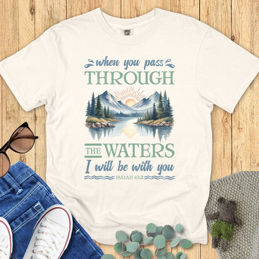 Comfort Colors Christian T-shirt in ivory with Isaiah 43:2 scripture design. Graphic shows a serene mountain and lake scene with sunrise and trees, surrounded by the verse “When you pass through the waters I will be with you.” Styled flat lay with denim jeans, sneakers, sunglasses, and greenery