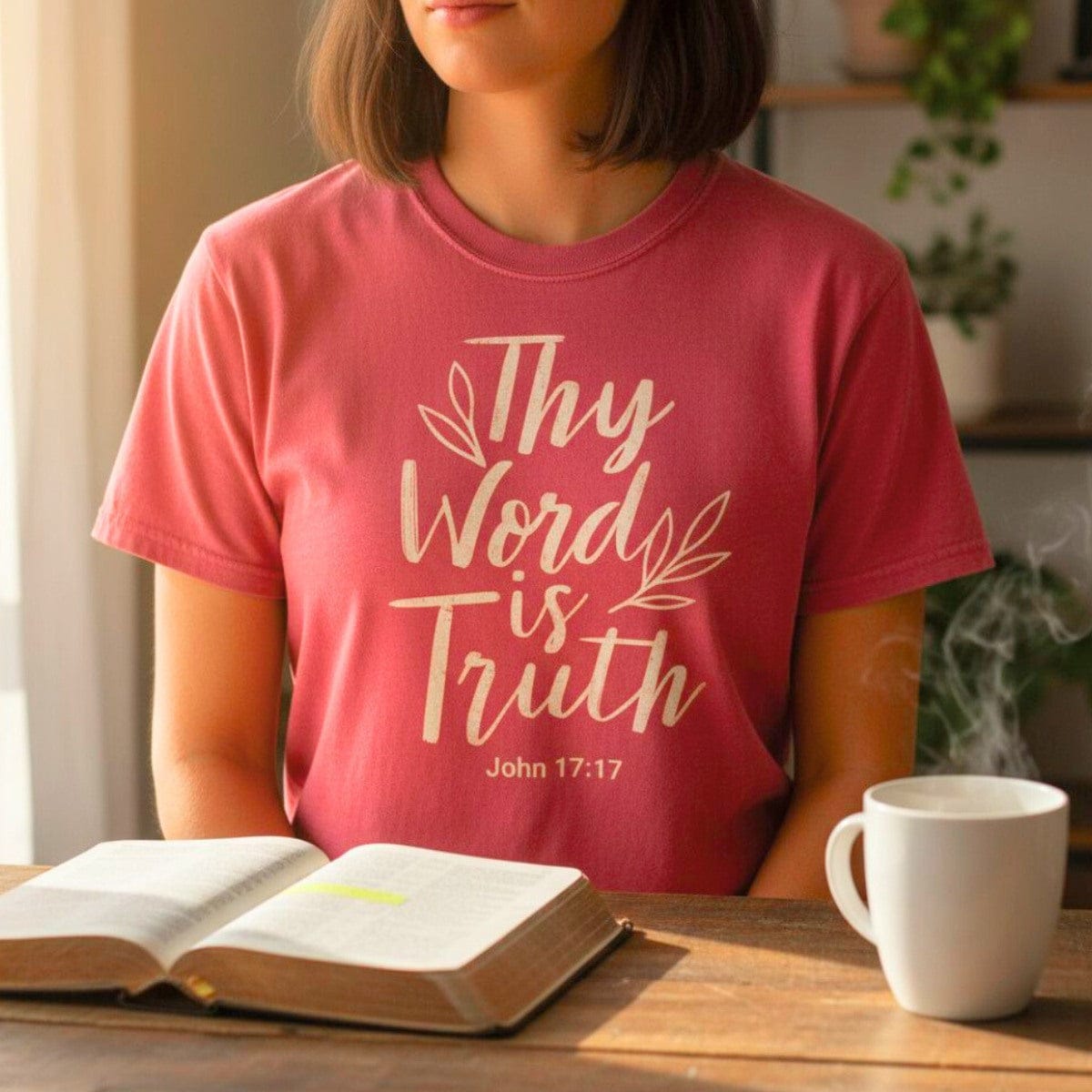 Woman sitting by a Bible and coffee cup wearing a Comfort Colors 1717 crimson “Thy Word Is Truth – John 17:17” t-shirt with elegant lettering. A cozy women’s scripture tee — meaningful faith-based apparel for women and modern Christian shirt, ideal Christian mom gift idea.
