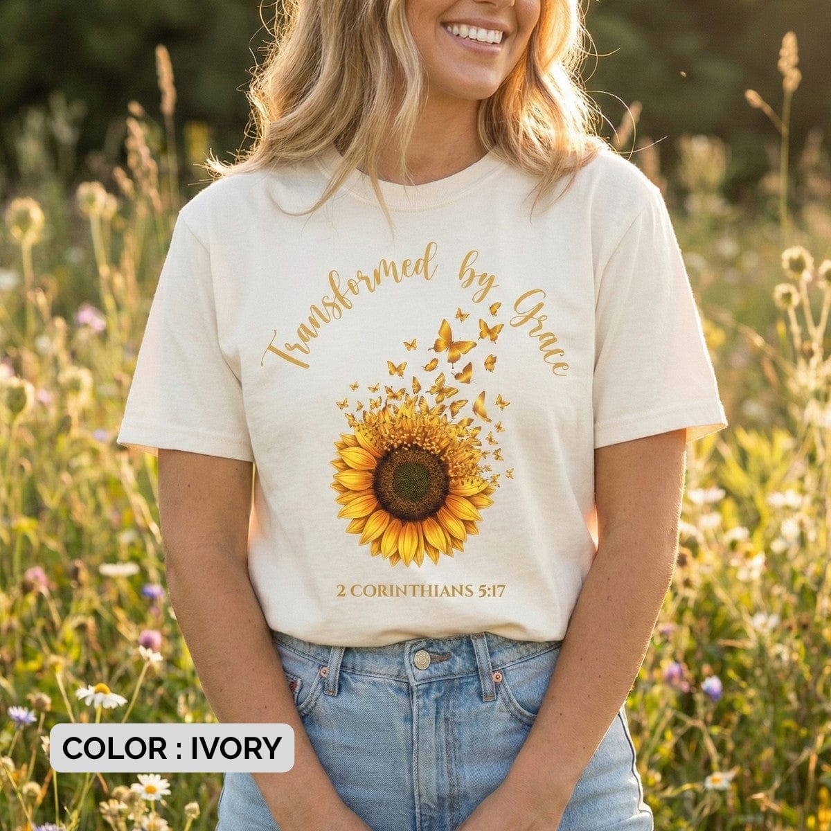 Ivory Bible Verse Shirt featuring a detailed graphic of a sunflower dissolving into flying butterflies, with the text "Transformed by Grace" and "2 Corinthians 5:17" in golden script.