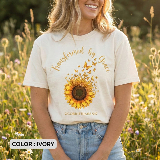 Ivory Bible Verse Shirt featuring a detailed graphic of a sunflower dissolving into flying butterflies, with the text "Transformed by Grace" and "2 Corinthians 5:17" in golden script.