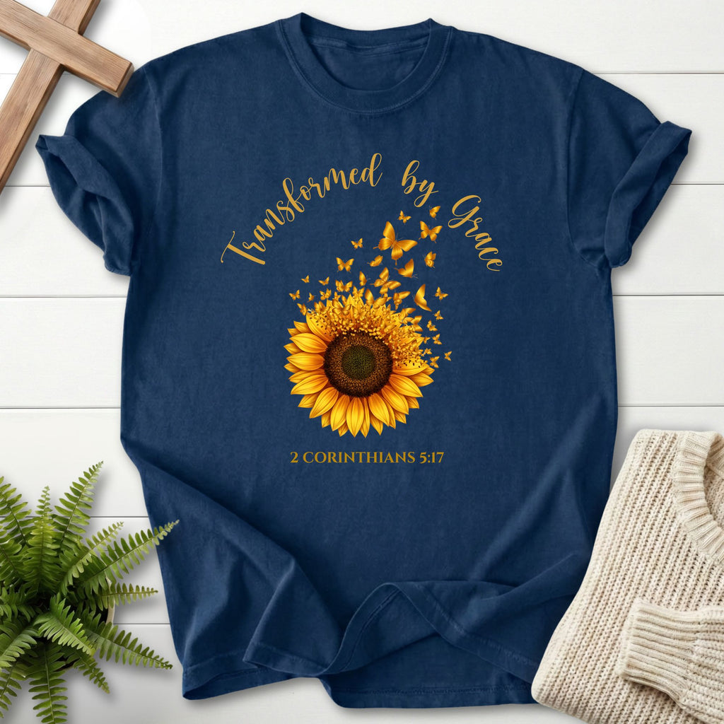 A navy blue graphic t-shirt laid flat on a white wood background, decorated with a yellow sunflower, butterflies, and the text 'Transformed by Grace' and '2 CORINTHIANS 5:17'. A wooden cross, a green fern, and a folded cream sweater are styled around the shirt.