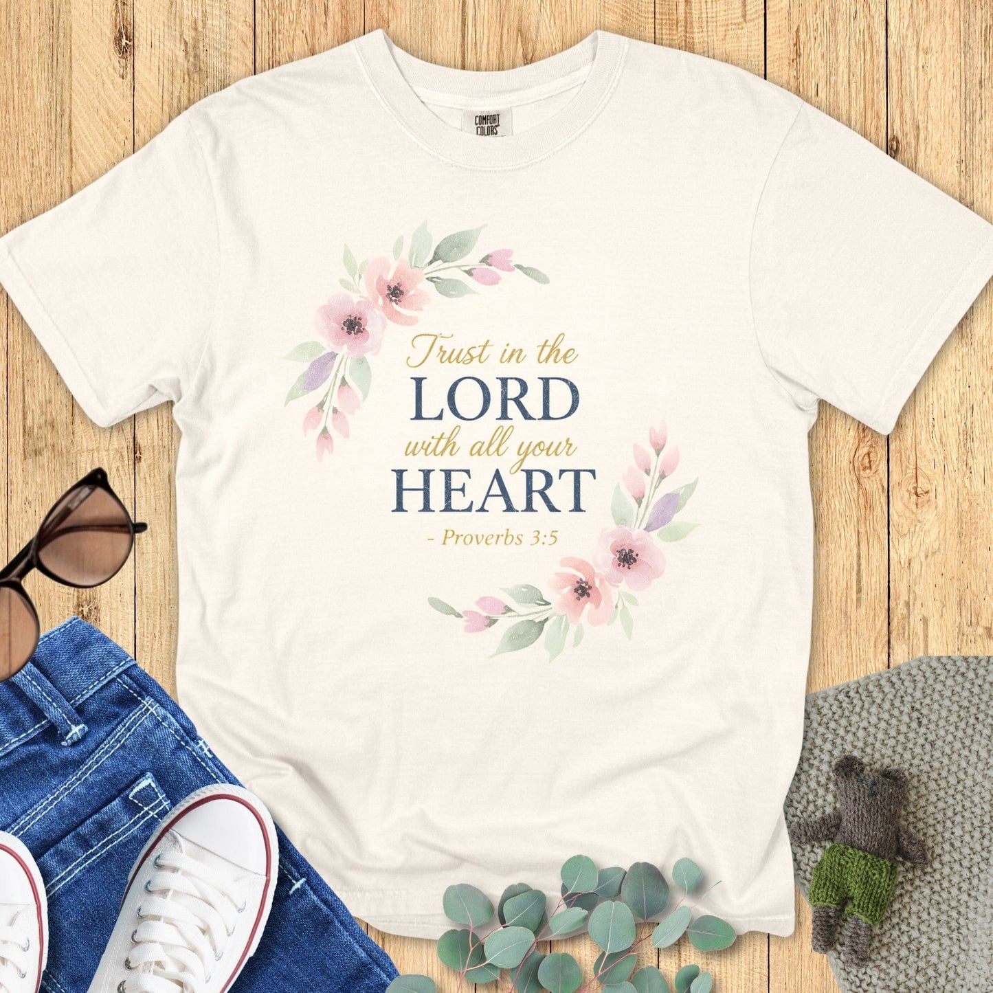 Flat lay of an ivory Comfort Colors Christian t-shirt featuring watercolor floral artwork around scripture text “Trust in the Lord with all your heart – Proverbs 3:5,” paired with denim and casual accessories