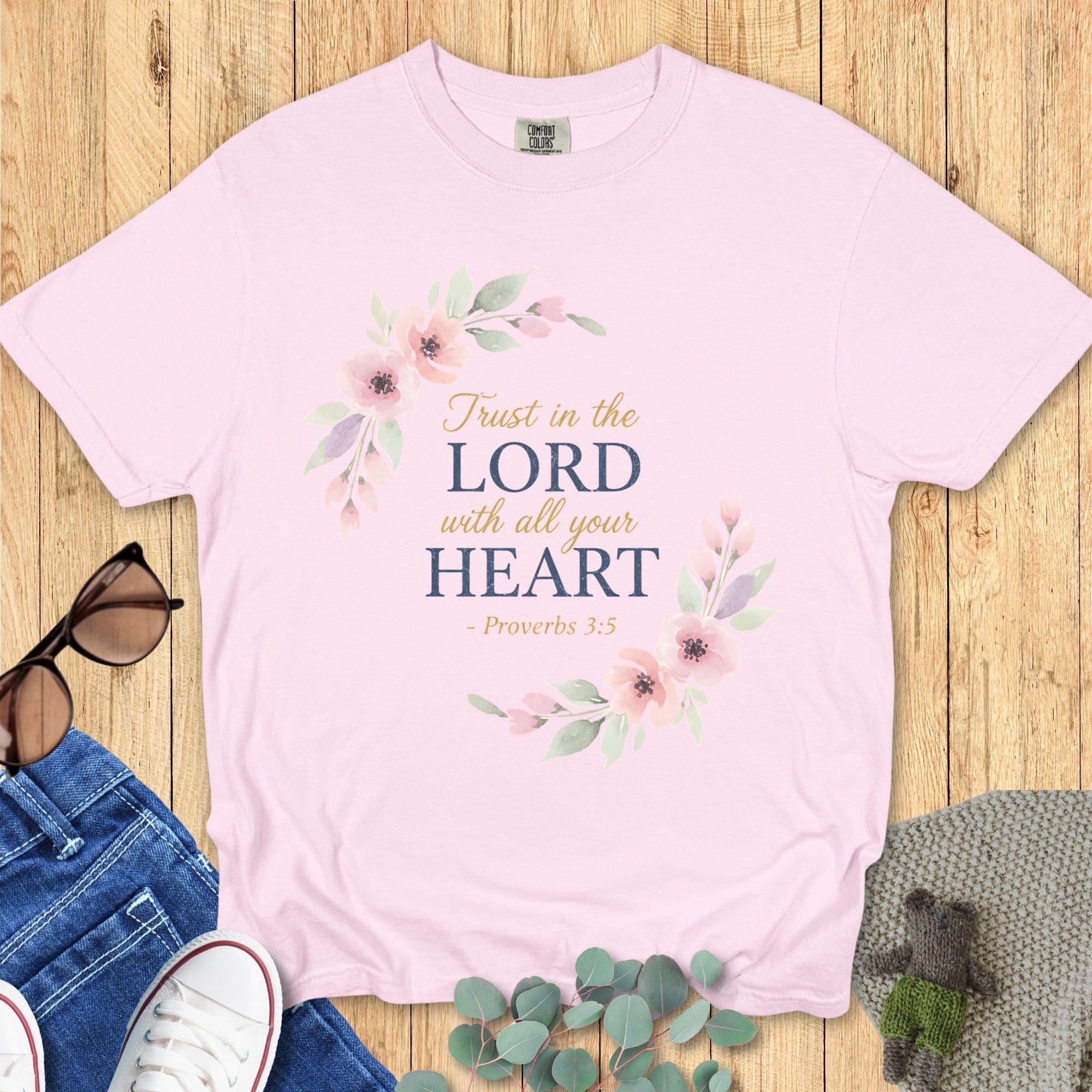 Flat lay of a light pink Comfort Colors Christian t-shirt with floral wreath design and Bible verse “Trust in the Lord with all your heart – Proverbs 3:5,” styled with jeans, sneakers, and sunglasses