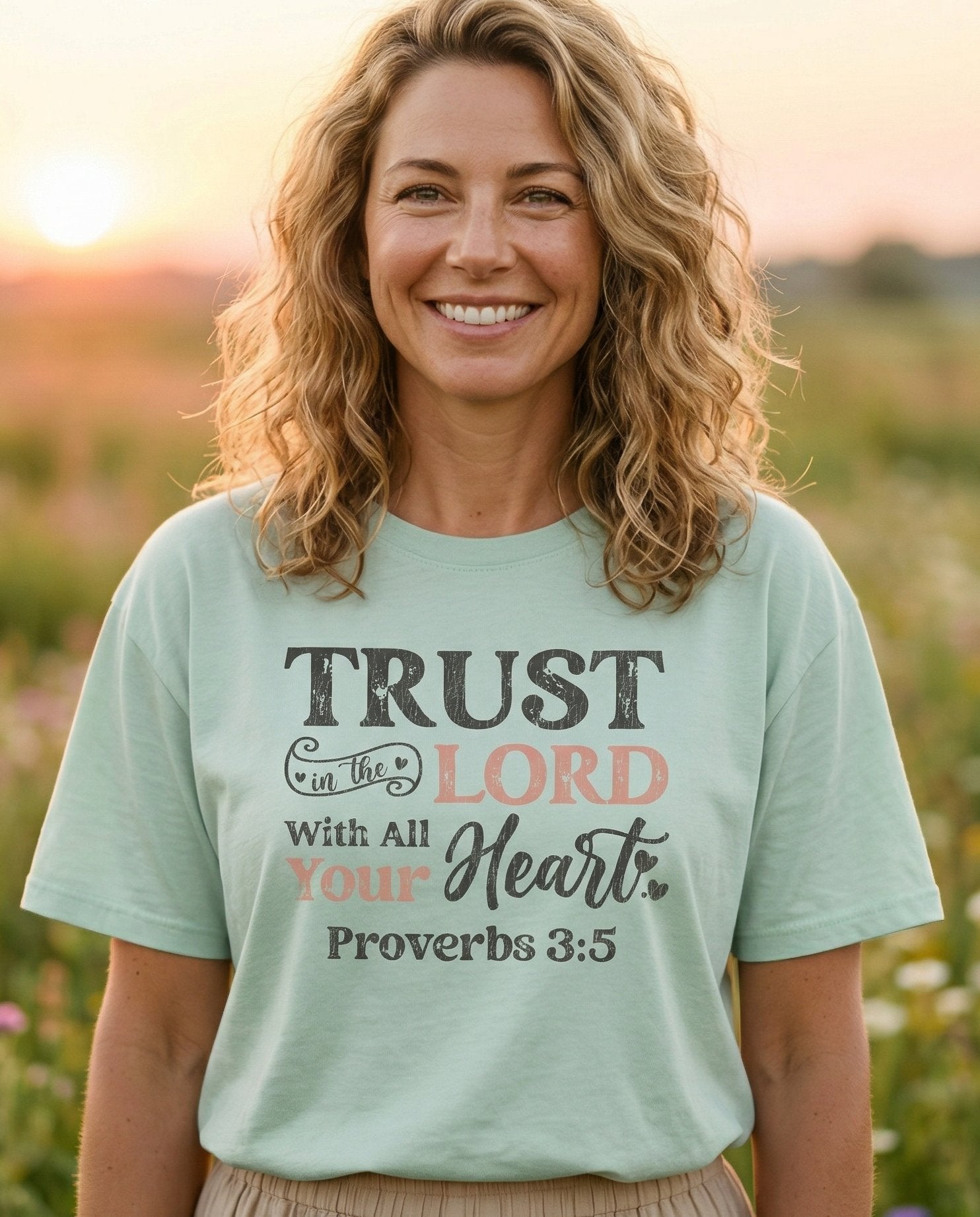 Trust in the Lord – Bay women's Christian tee, lifestyle photo in wildflower meadow at sunrise – Bold distressed mixed-font design with 'Trust in the LORD With All You – Proverbs 3:5 – She Wears Faith