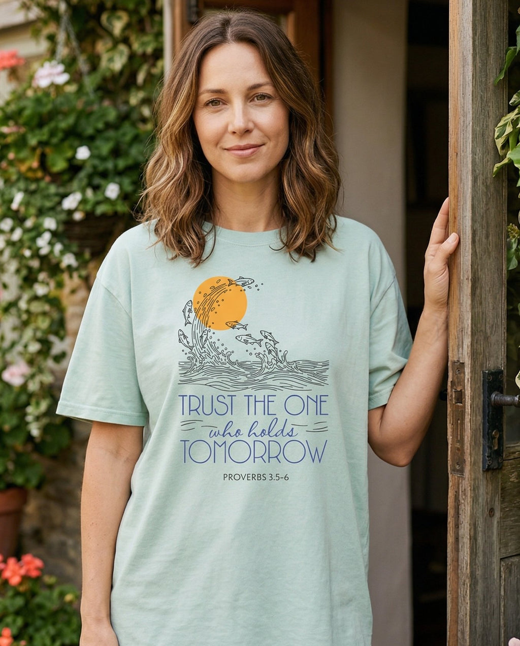 Trust the One who holds Tomorrow – Bay women's Christian tee, lifestyle photo in cottage doorway – Line-art illustration of fish leaping from ocean waves around a golden – Proverbs 3:5-6 – She Wears Faith