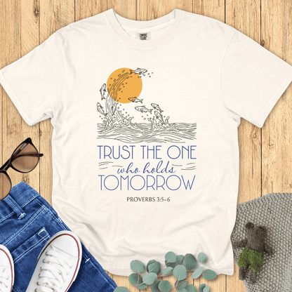 Flat lay of an ivory Comfort Colors Christian t-shirt with the Bible verse Proverbs 3:5-6 design. The shirt features a hand-drawn wave and fish graphic under a golden sun with the words “Trust the One Who Holds Tomorrow”. Styled with denim jeans, white sneakers, and sunglasses on a wooden background