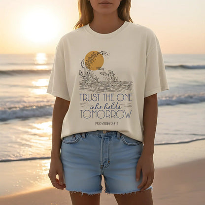 Woman wearing a natural ivory Comfort Colors Christian t-shirt on the beach at sunset. The Proverbs 3:5-6 design shows waves, fish, and a golden sun, paired with the words “Trust the One Who Holds Tomorrow”. Styled with denim shorts, creating a relaxed faith-inspired look