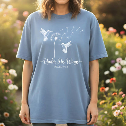 Blue jean Comfort Colors Christian T-shirt with Psalm 91:4 verse "Under His Wings." Features white hummingbird and dandelion design. Modeled outdoors in a flower garden setting