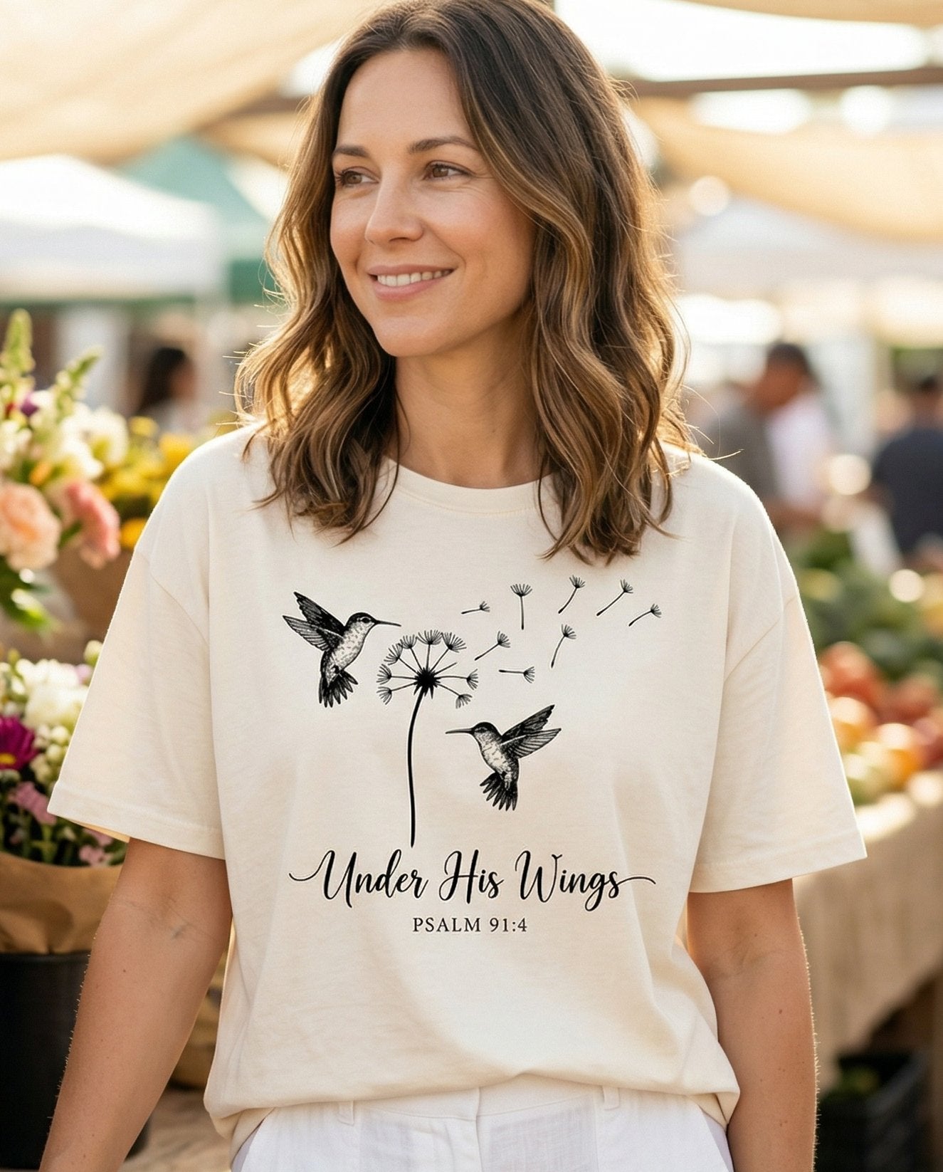 Under His Wings – Ivory women's Christian tee, lifestyle photo in farmers market – Delicate line art of two hummingbirds flanking a dandelion in full see – Psalm 91:4 – She Wears Faith